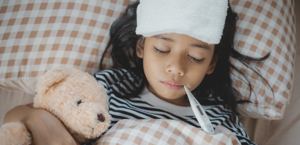 طفلة مريضة على رأسها قطعة قماش وفي فمها ميزان حرارة وبجانبها لعبتها