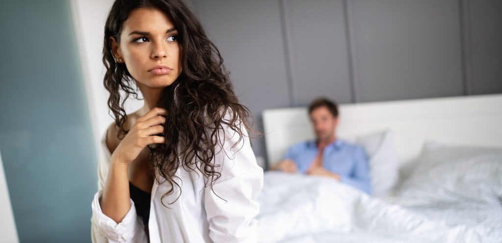 امرأة تجلس على طرف السرير وخلفها زوجها يجلس فيه