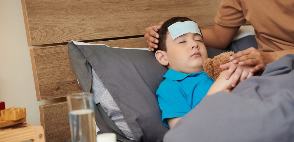 أمّ تجلس إلى جانب طفلها النائم على السرير وعلى رأسه كمّادة