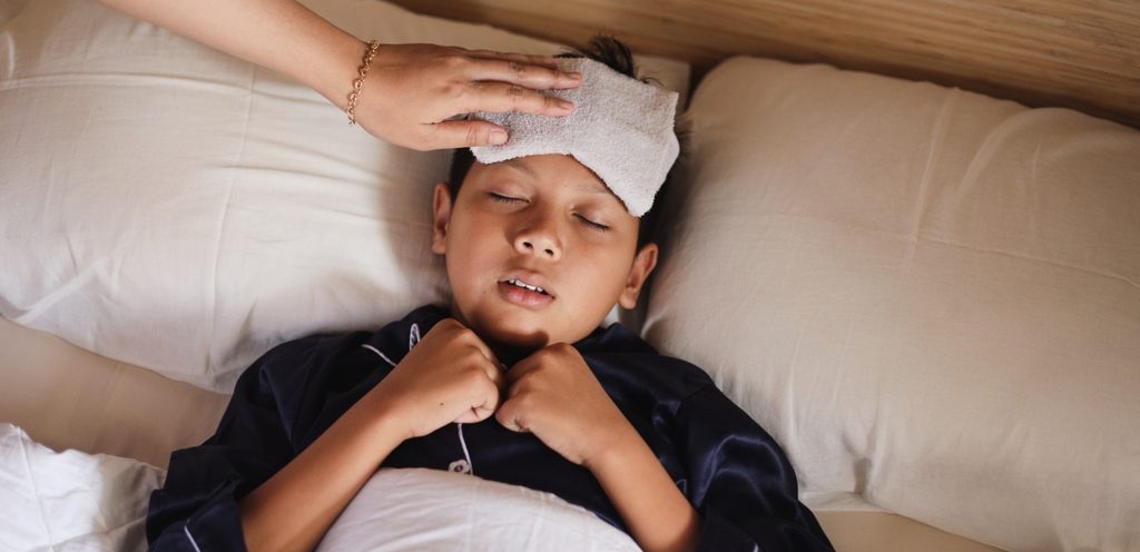 طفل نائم مغطّى في السرير ووالدته تضع قطعة من القماش على رأسه لخفض حرارته