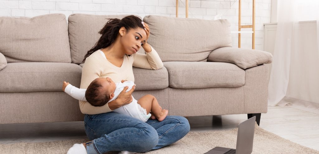 أمّ متعبة تجلس على الأرض تحمل طفلها وهو يبكي وأمامها حاسوب محمول