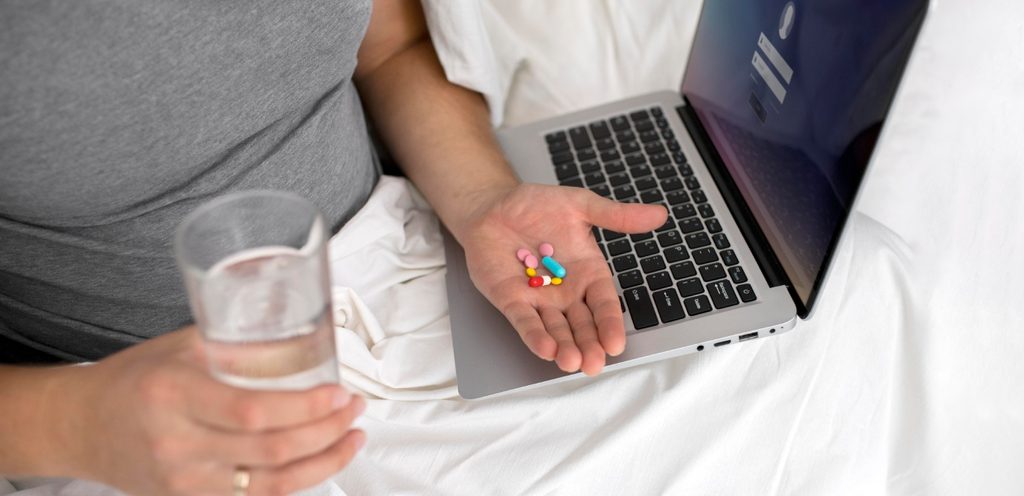 امرأة تجلس في سريرها تمسك في يدها مجموعة من حبوب الأدوية وكوب من الماء في اليد الأخرى وأمامها حاسوب محمول