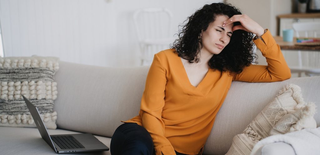 امرأة تجلس على الكنبة تغلق عينيها بشدّة وتضع يدها على رأسها وبجانبها حاسوب محمول