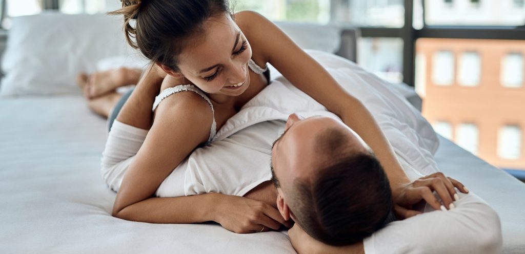 زوج مستلقٍ على ظهره على السرير وزوجته مستلقية فوقه عليه وهما ينظران إلى بعضهما البعض بسعادة ومبتسمان