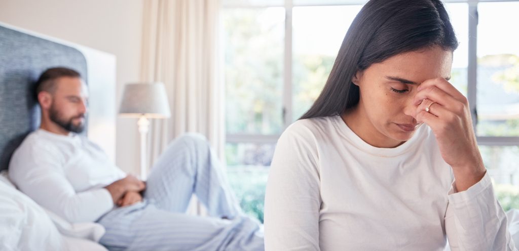امرأة حزينة تضع أصابعها بين عينيها وخلفها زوجها يجلس على السرير
