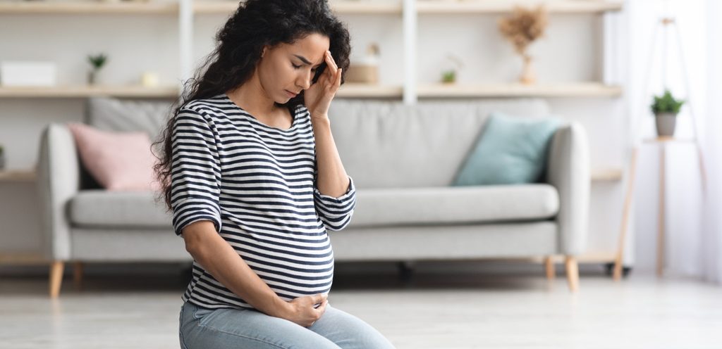 امرأة حامل حزينة تجلس على الأرض وتضع يدًا على رأسها والأخرى على بطنها