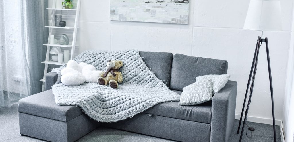 أريكة باللون الرمادي الغامق عليها غطاء باللون الرمادي الفاتح ودمية