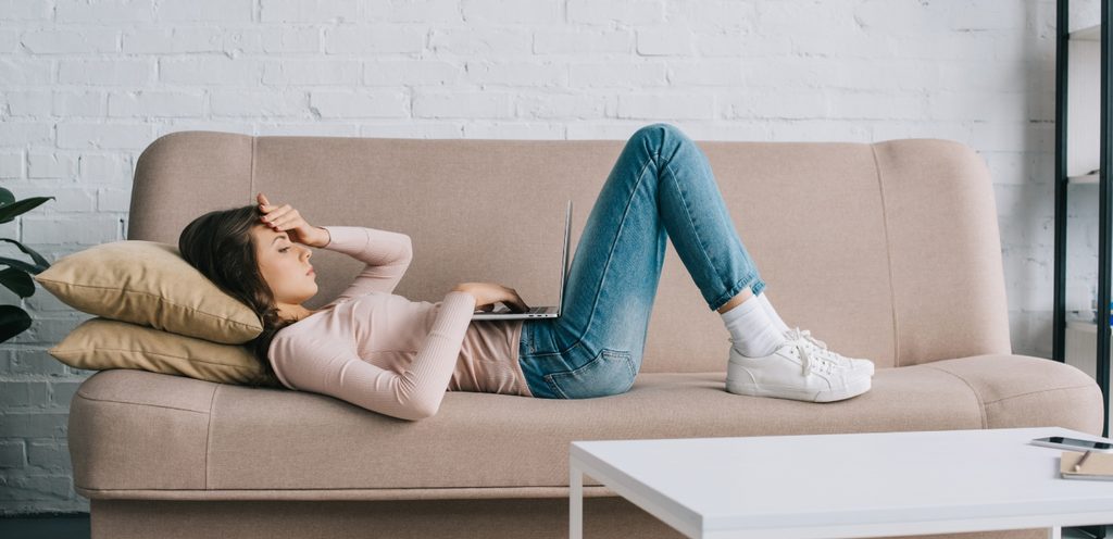 امرأة مستلقية على ظهرها على الكنبة تضع يدها على رأسها وعلى بطنها حاسوب محمول