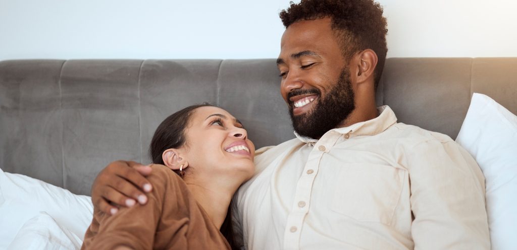 زوج يعانق زوجته وهما يجلسان على السرير وينظران إلى بعضهما البعض ويبتسمان