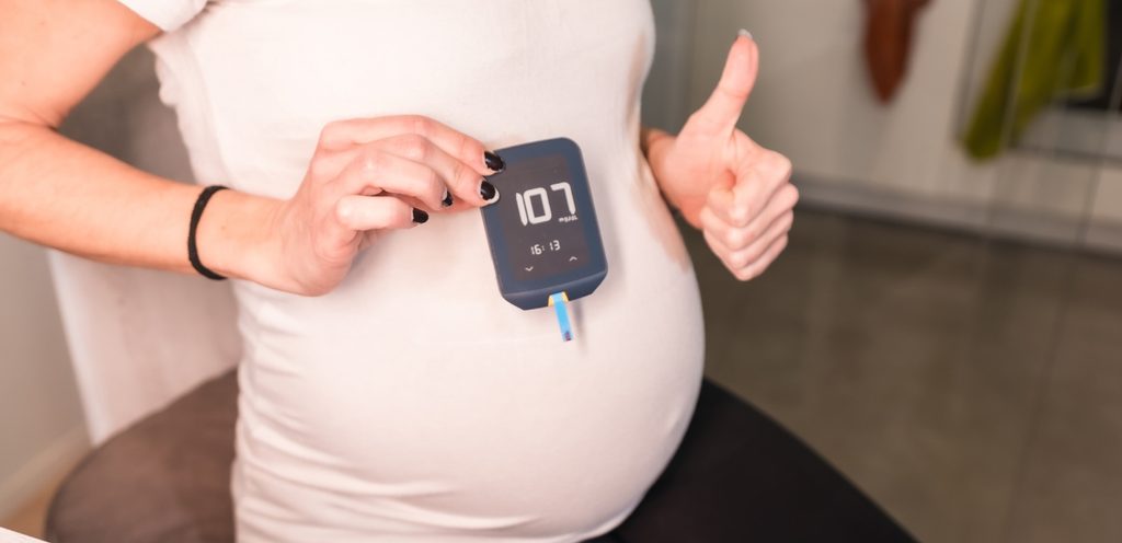 امراة حامل تحمل جهاز سكّر الحمل أمام بطنها وترفع إبهامها في اليد الأخرى