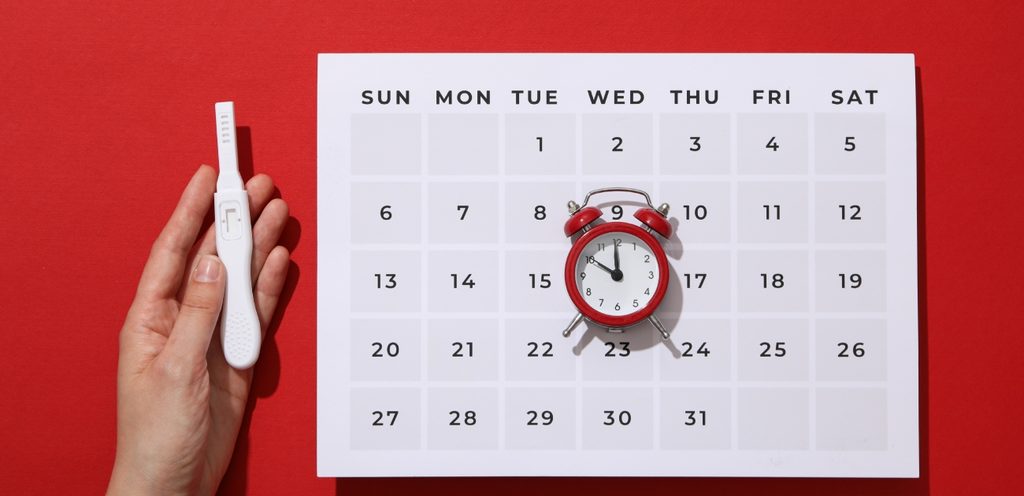 يد امرأة تمسك اختبار حمل وبجانبها ورقة تواريخ ومنبّه باللون الأحمر