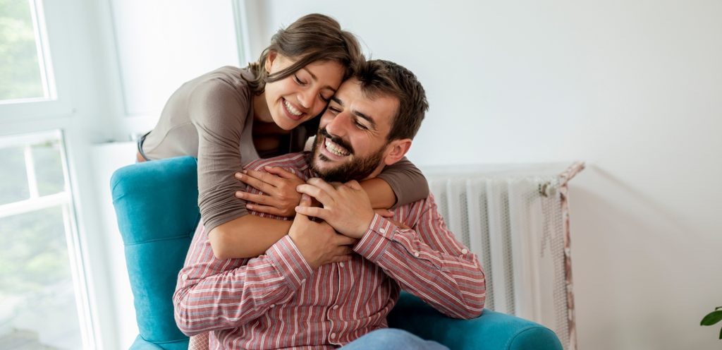 رجل يجلس على الكنبة وزوجته تُعانقه من الخلف وهما يبتسمان