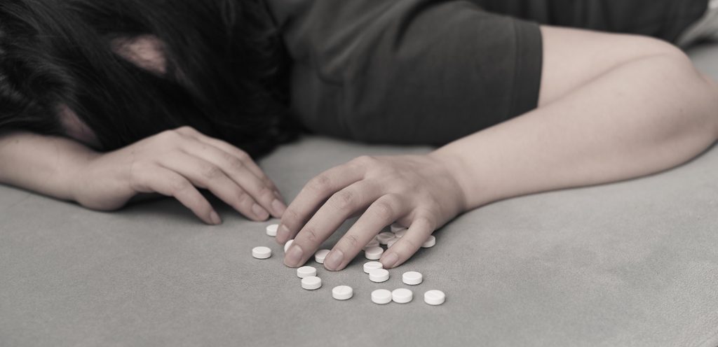 امرأة تحني رأسها وتسنده على السرير وأسفل يدها حبوب من الدواء