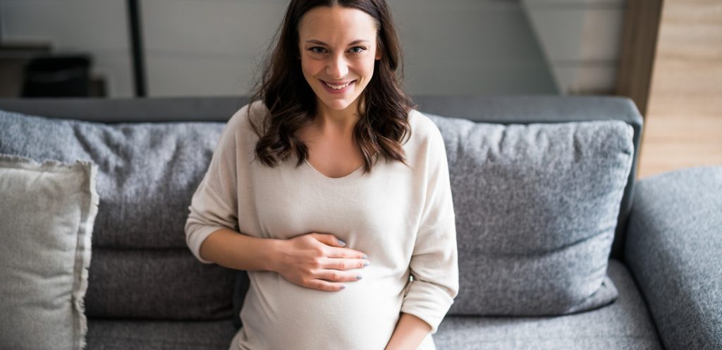 امرأة حامل تجلس على الكنبة وتضع يديها على بطنها وهي مُبتسِمة