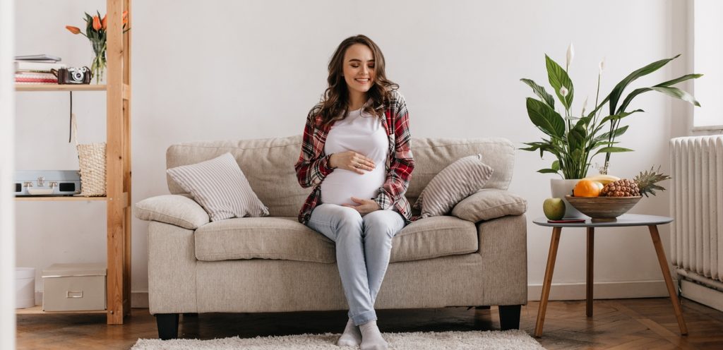 امرأة حامل تجلس على الكنبة وتضع يديها على بطنها وتبتسم