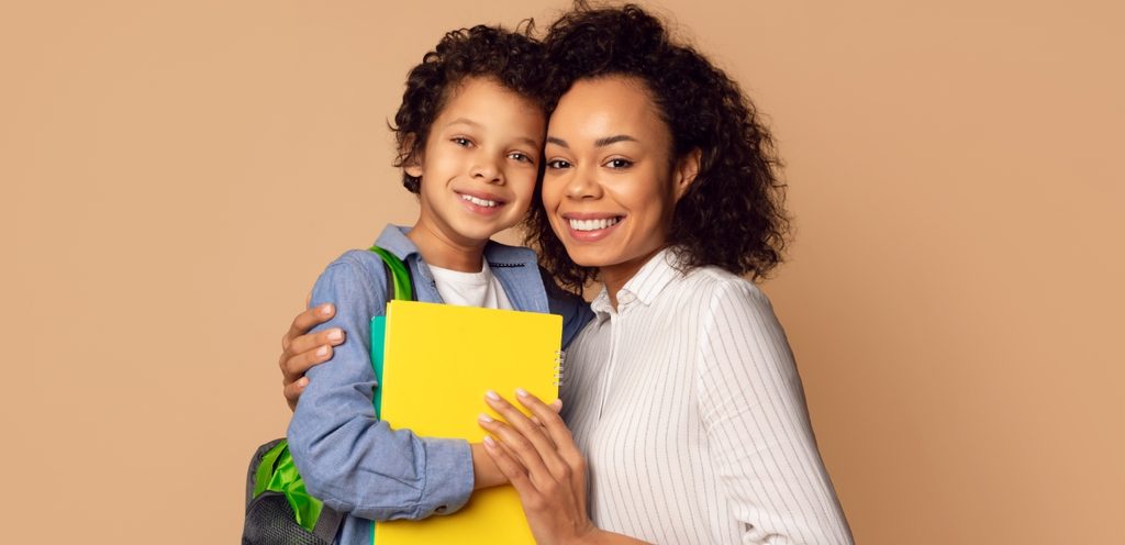 أمّ وطفلها يمسك كتابًأ ويضع حقيبة مدرسيّة على ظهره يتعانقان جنبًا إلى جنب ويبتسمان