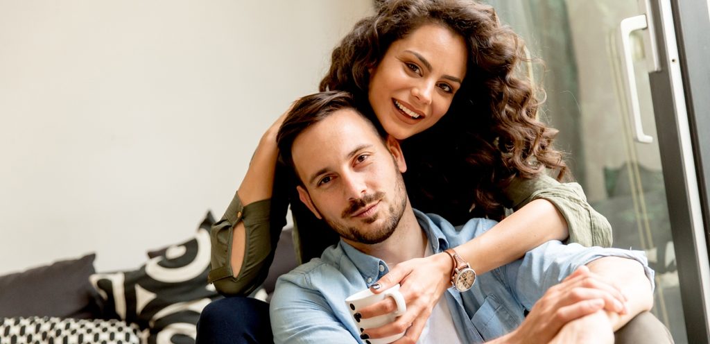 امرأة تُعانق زوجها من الخلف وتضع يدها حول رقبته مبتسمة