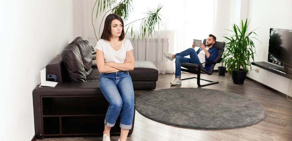 امرأة حزينة تجلس على طرف الكنبة وخلفها رجل يجلس على كرسي ويمسك جهاز لوحي