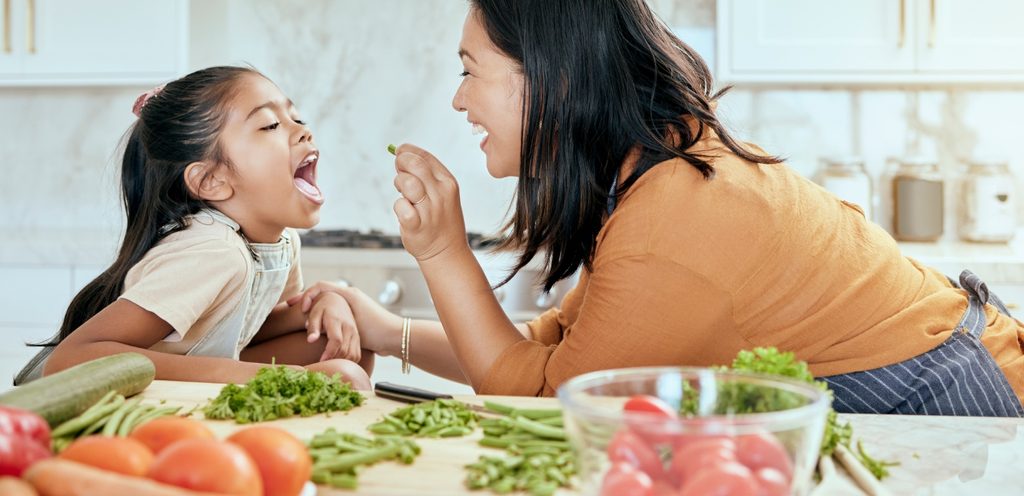 أمّ تُطعِم طفلتها قطعة خضار صغيرة وأمامهما طماطم وخس وخيار