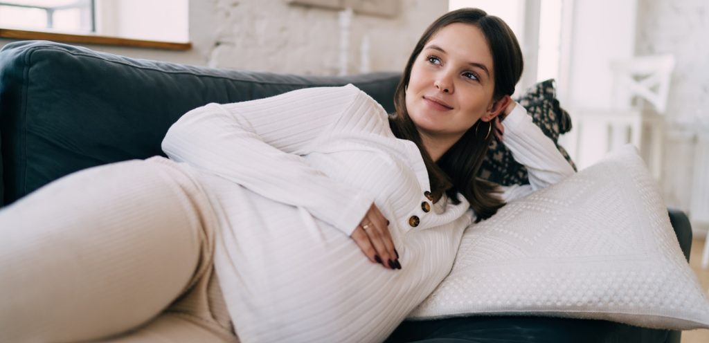امرأة حامل مستلقية على الكنبة تضع يدها على بطنها ومبتسمة