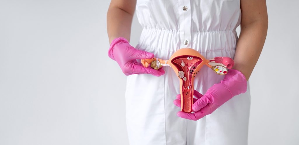 امرأة تلبس رداءً باللون الأبيض وفي يديها كفوف زهري تُمسِك مجسّم العضو التناسلي الأنثوي