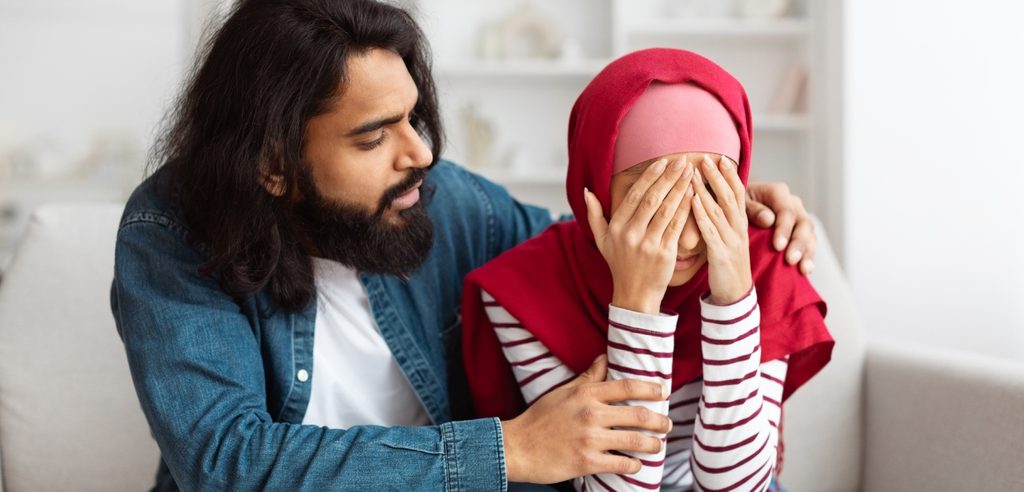 امرأة عربيّة محجّبة حزينة تضع يديها على وجهها وزوجها يواسيها