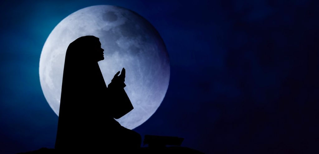 امرأة مسلمة ترفع يديها للدعاء والقمر خلفها
