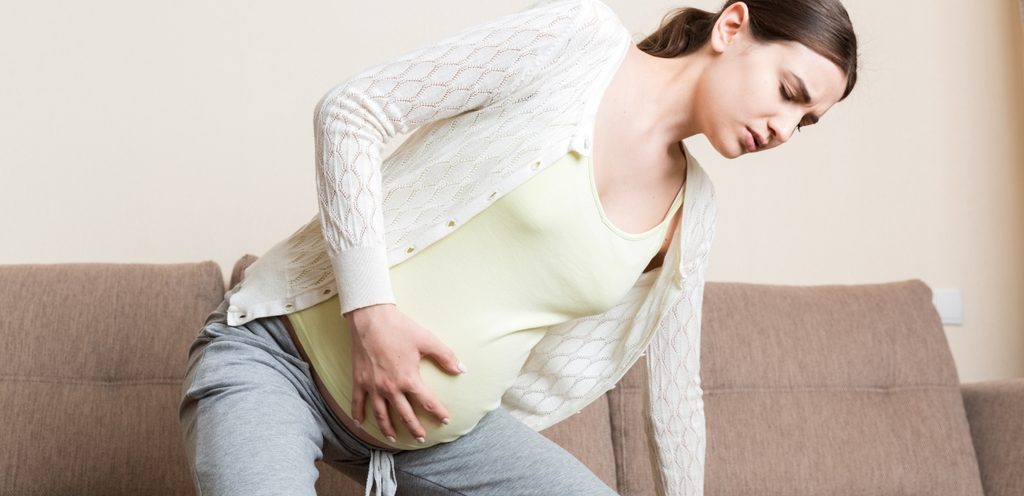 امرأة حامل تبدو غير مرتاحة تضع يدها على بطنها وتحاول الجلوس على الكنبة