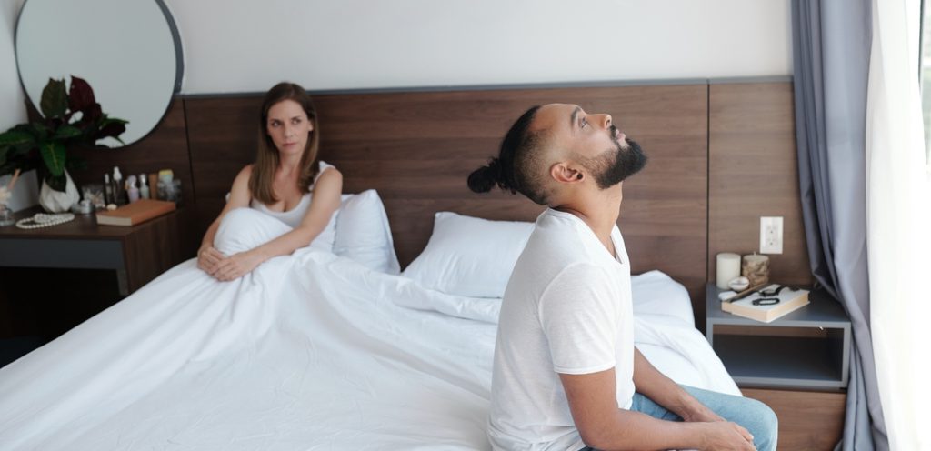 امرأة حزينة في السرير وزوجها يجلس على طرف الفراش ويرفع رأسه لأعلى