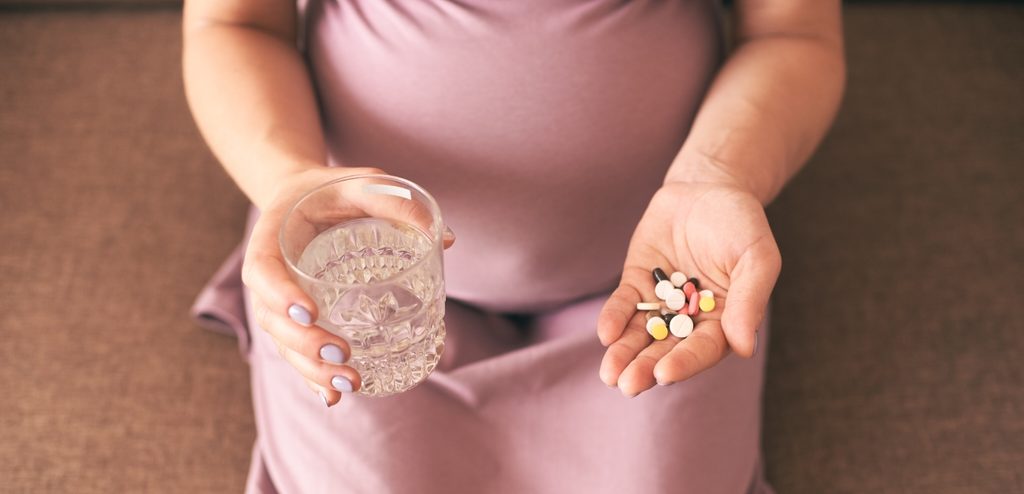 امرأة حامل تمسك كوبًا من الماء وحبوبًا من الدواء