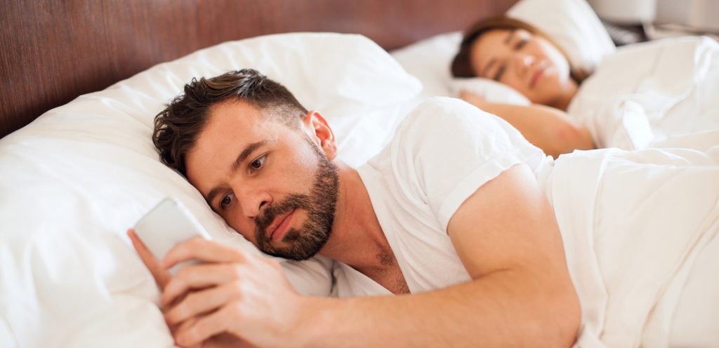 رجل يمسك هاتفه في الفراش وزوجته بجانبه