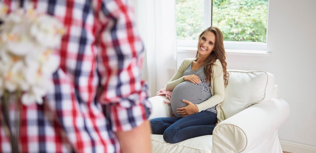 امرأة حامل سعيدة تنظر إلى زوجها الذي يحمل الورد خلف ظهره