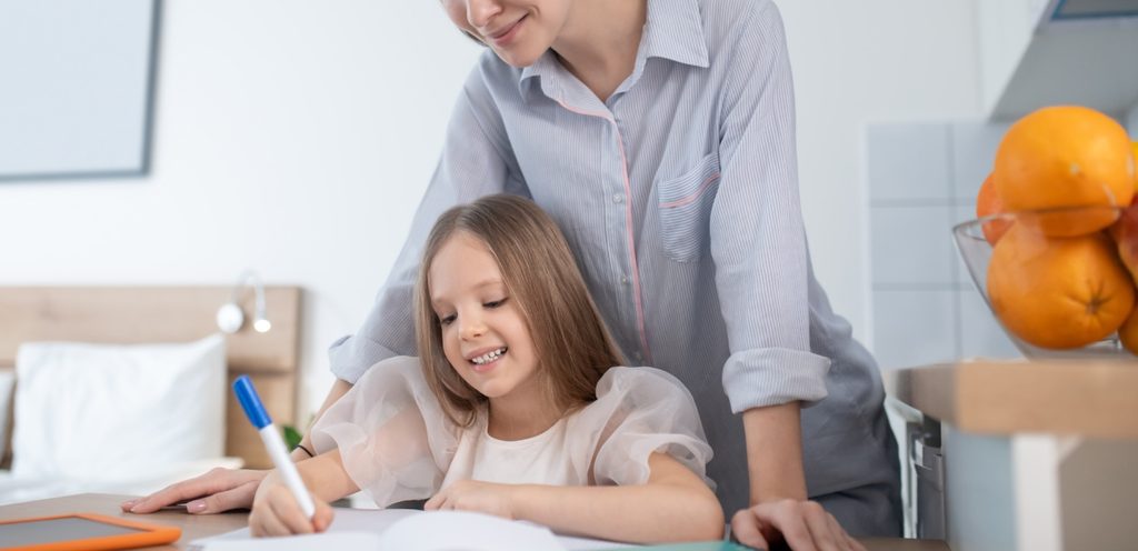طفلة تحلّ واجباتها المدرسيّة ووالدتها تقف خلفها