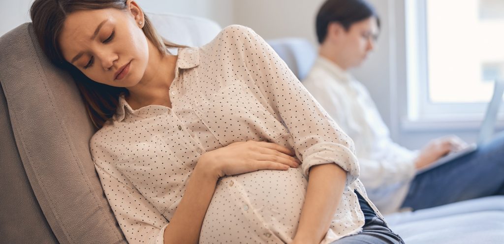 امرأة حامل حزينة وزوجها يجلس خلفها يعمل على الحاسوب