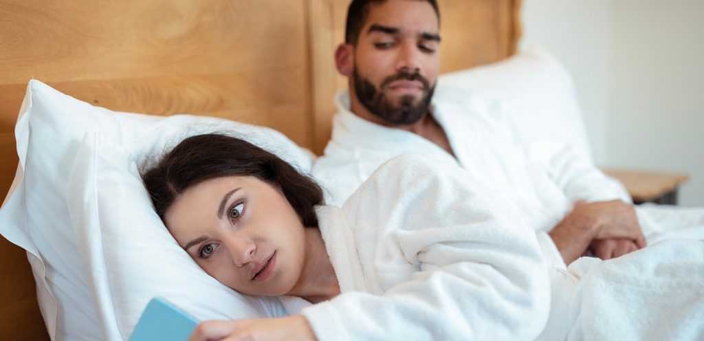 امرأة تمسك هاتفها وزوجها بجانبها على السرير ينظر إليها