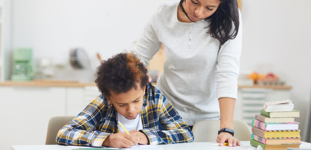 امرأة تقف بجانب طفلها وهو يحلّ واجباته المدرسيّة