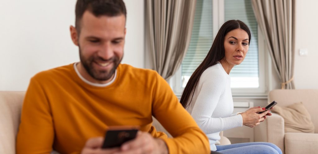 امرأة تنظر إلى زوجها الذي يستخدم هاتفه ويضحك